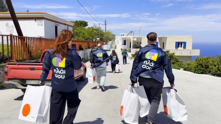 Μέλη της ομάδας CSR 'πλάι σου' του Plaisio, με τζάκετ που φέρουν το λογότυπο, περπατούν σε δρόμο νησιωτικού οικισμού κρατώντας τσάντες με σχολικά είδη, σε αποστολή διανομής σε παιδιά δημοτικού στη Σέριφο