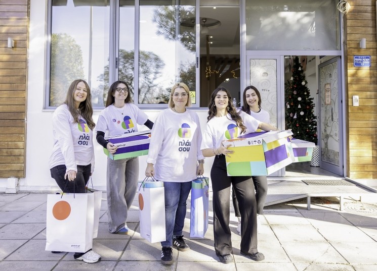 Πέντε γυναίκες μέλη της CSR ομάδας 'πλάι σου' του Πλαισίου, φορώντας μπλουζάκια με το λογότυπο, κρατούν έγχρωμα δώρα και τσάντες μπροστά σε κτίριο με χριστουγεννιάτικο δέντρο.