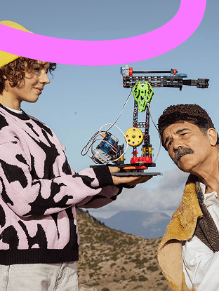 Νεαρή γυναίκα κρατά κατασκευή ρομπότ από LEGO-like κομμάτια, ενώ άνδρας δίπλα της την κοιτάζει έκπληκτος, σε εξωτερικό τοπίο με βουνά